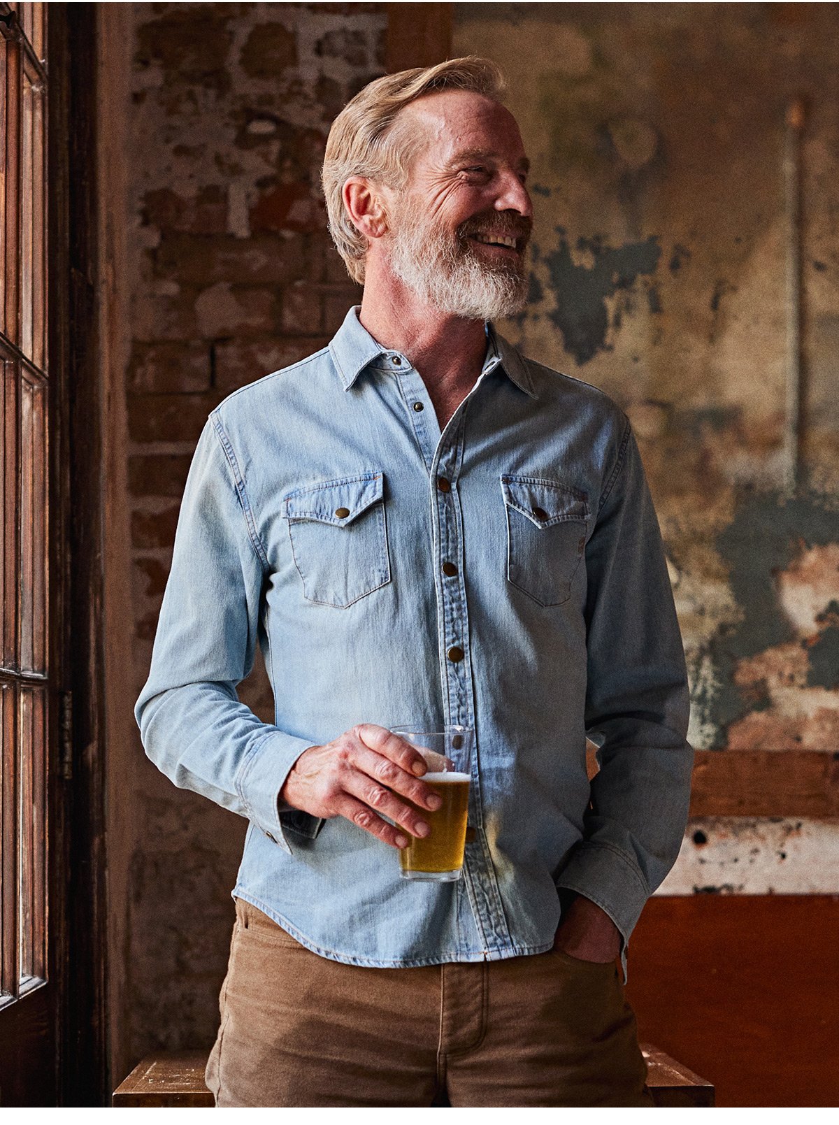 Male model wears the Shoals Denim Shirt in denim wash. He laughs while holding a beer. 